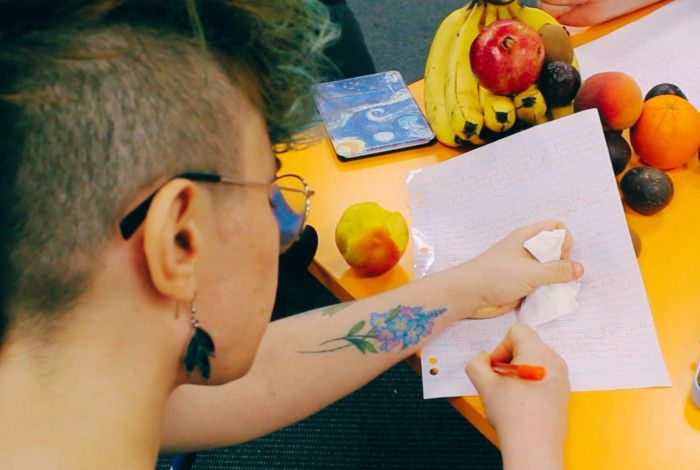 Person writing at desk with fruit and pens around.